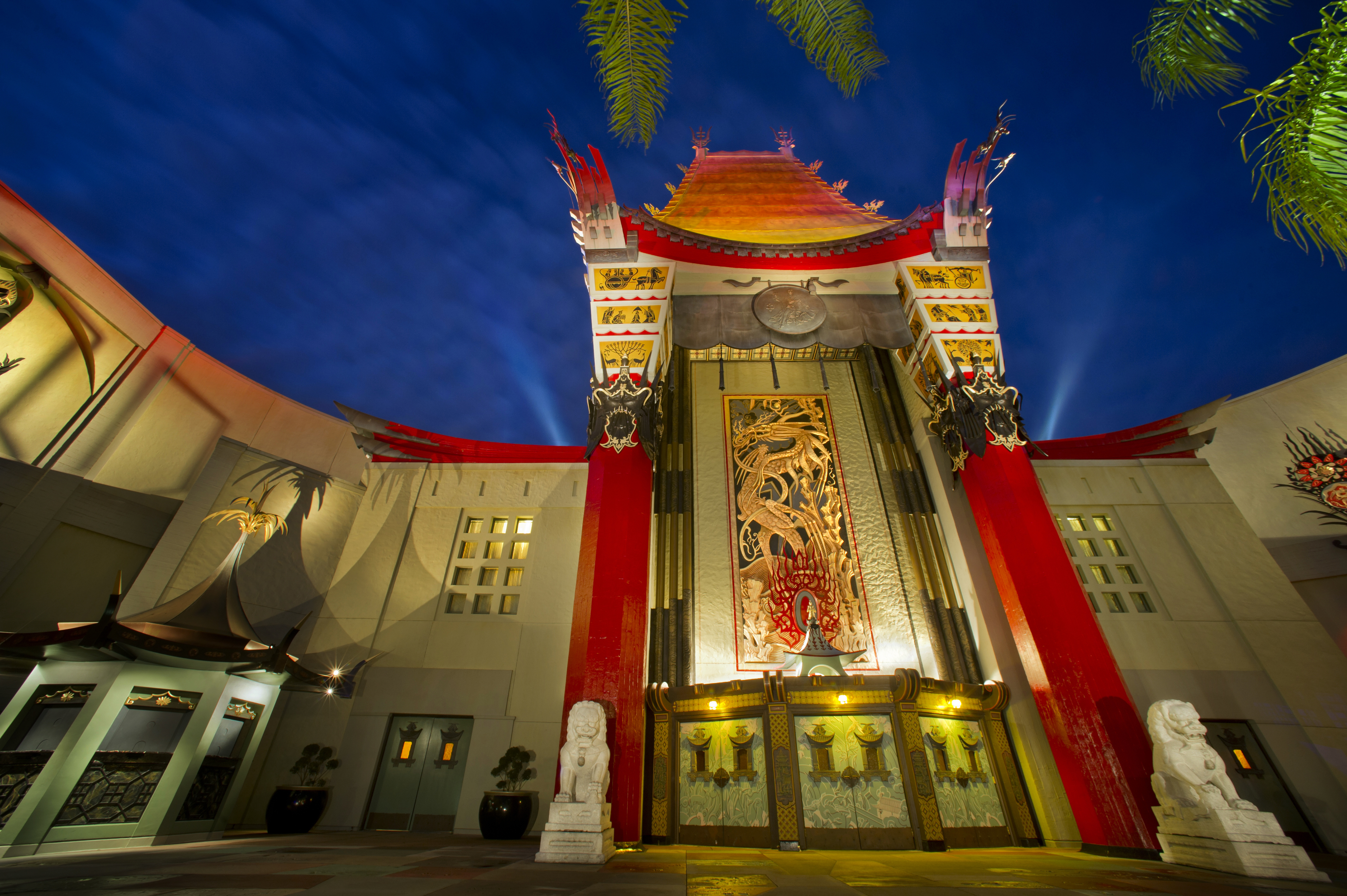 An up-looking angle of the Chinese Theater in Disney's Hollywood Studios at night with spotlights in the background and the red, orange, yellow and beige building itself lit up by lights.
