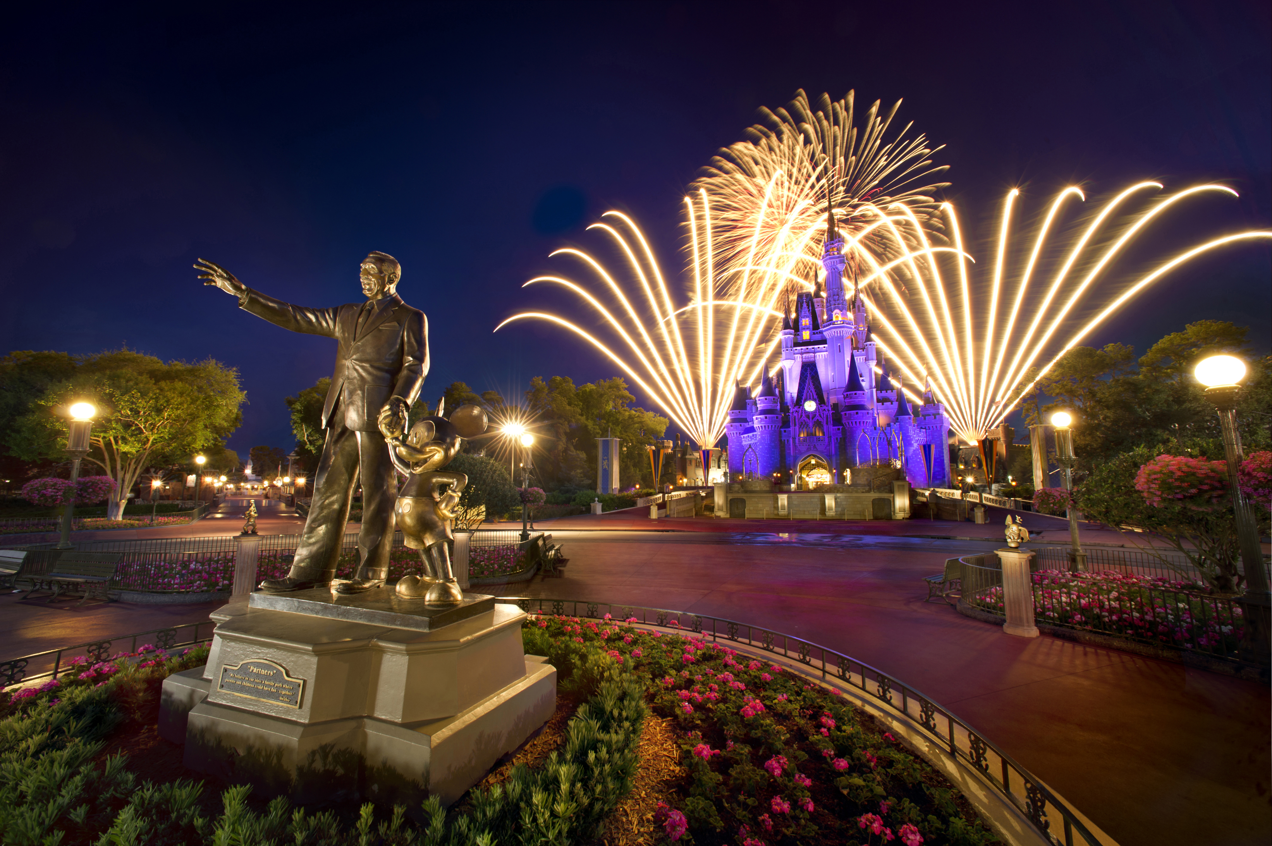 Nighttime view of gold Walt and Mickey statue in front of purple-lit Cinderella Castle with bright fireworks going off behind the castle at Disney's Magic Kingdom. Surrounding everything are trees and pink flowers in the landscaping.