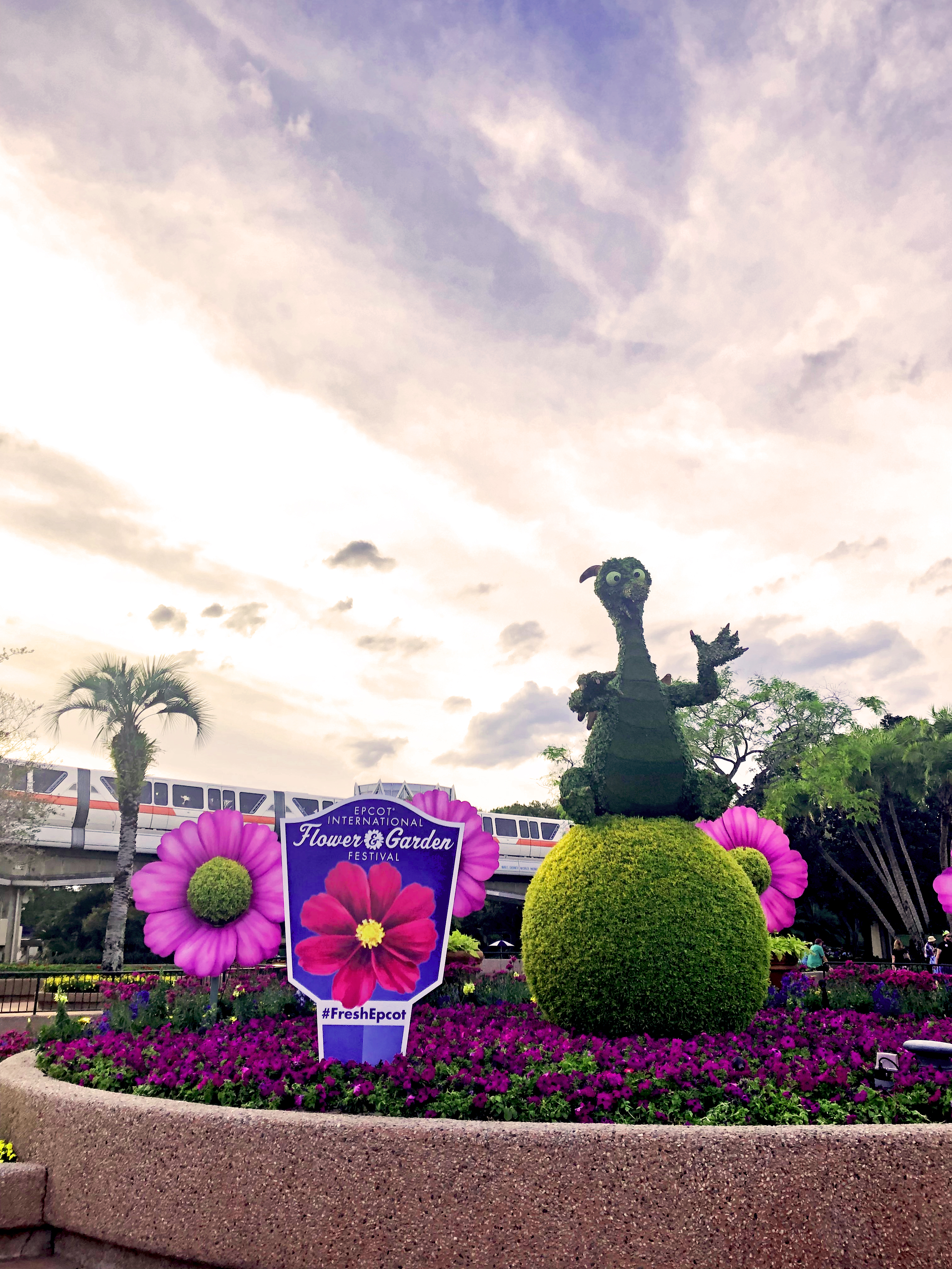 Daytime shot of Figment shrub sitting on top of rounded bush. Sign in front to the left of Figment that reads "Epcot International Flower & Garden Festival" colored in purple with a large pink flower on the sign and below that, labeled "#FreshEpcot". Everything is surrounded by massive purple flowers and small purple flowers with a orange-stripe monorail driving past in the background.