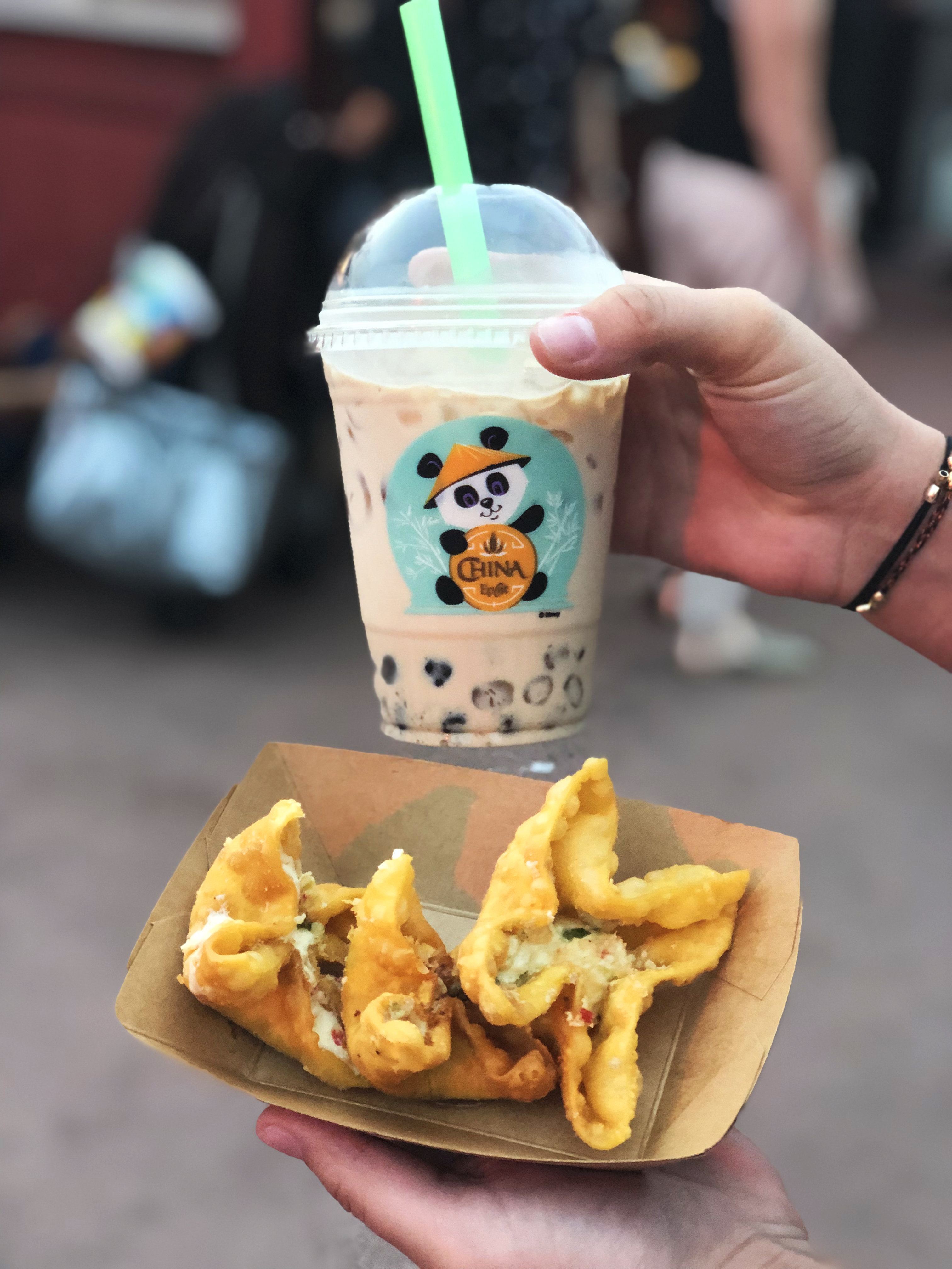 Photo of girls hands holding a cream-colored beverage with boba, a green straw and a panda on the cup (right hand), and a food boat of two stuffed wontons (left hand) during the daytime.