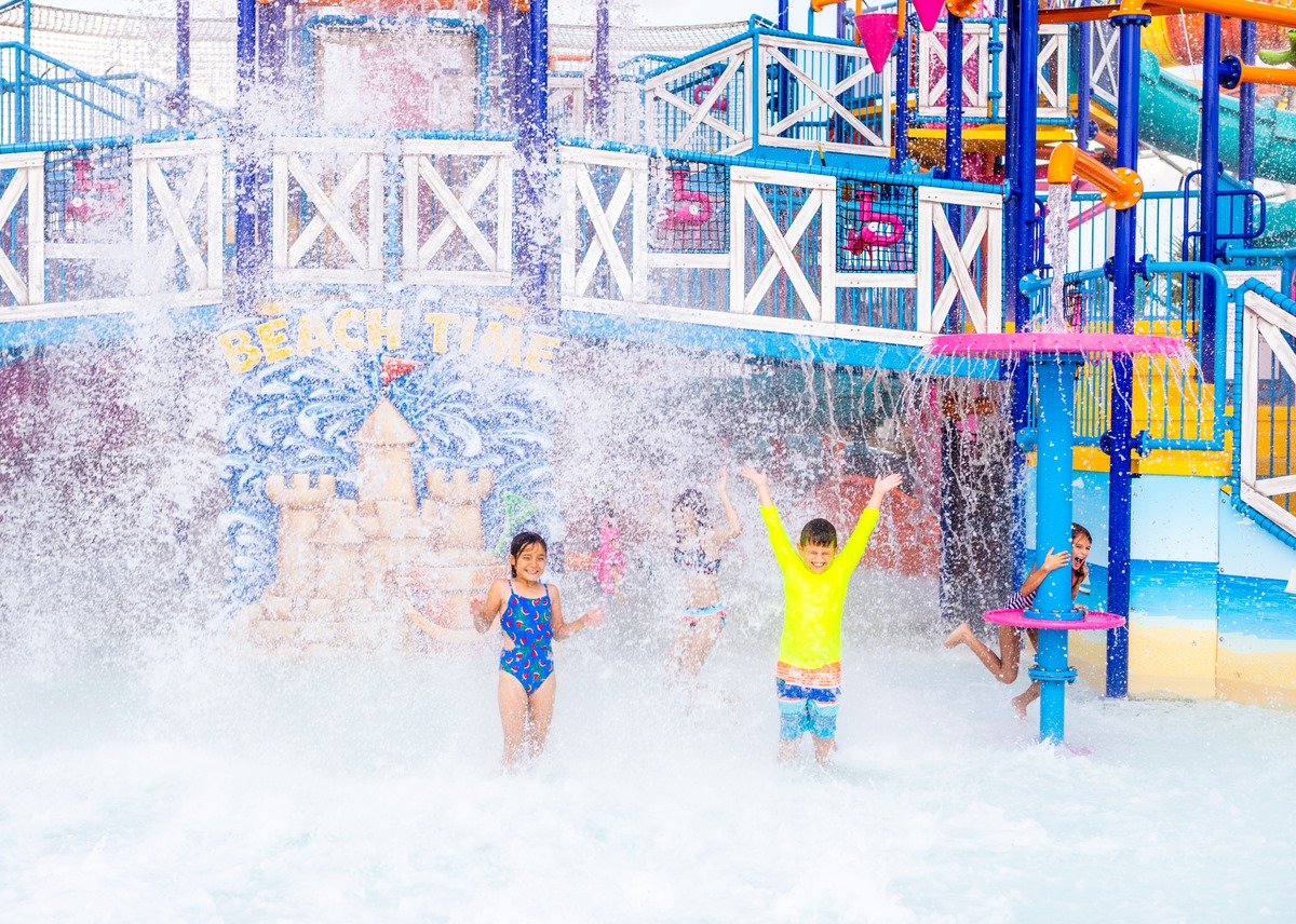 During daytime. Children with eyes closed and hands up in the air to touch the water splashing down on them at the kids area of Island H2O Live!'s water park.