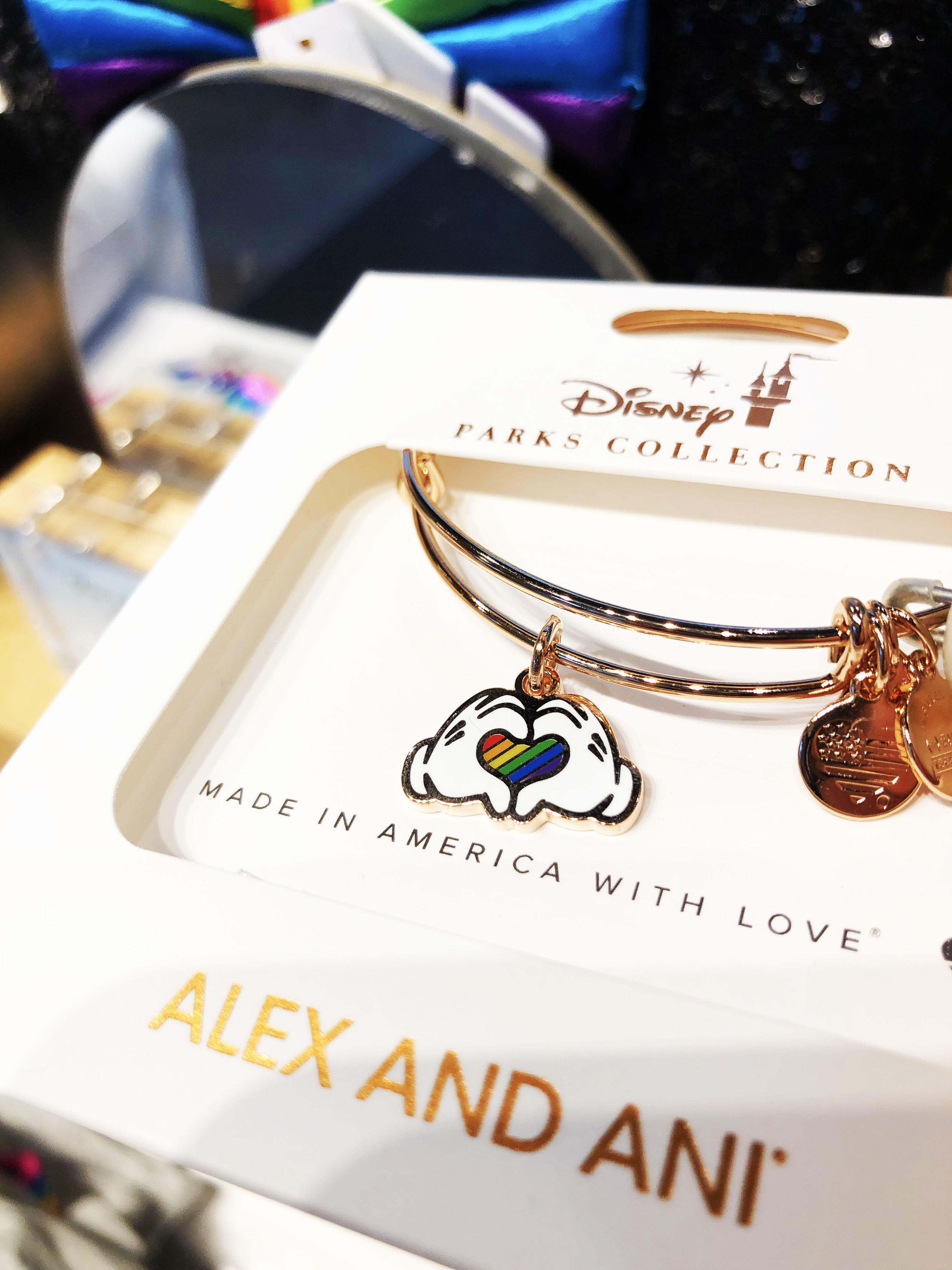 A right-angle shot of a packaged Alex and Ani bracelet. Bracelet is rose gold with a charm of Mickey hands making a heart that is rainbow-striped.