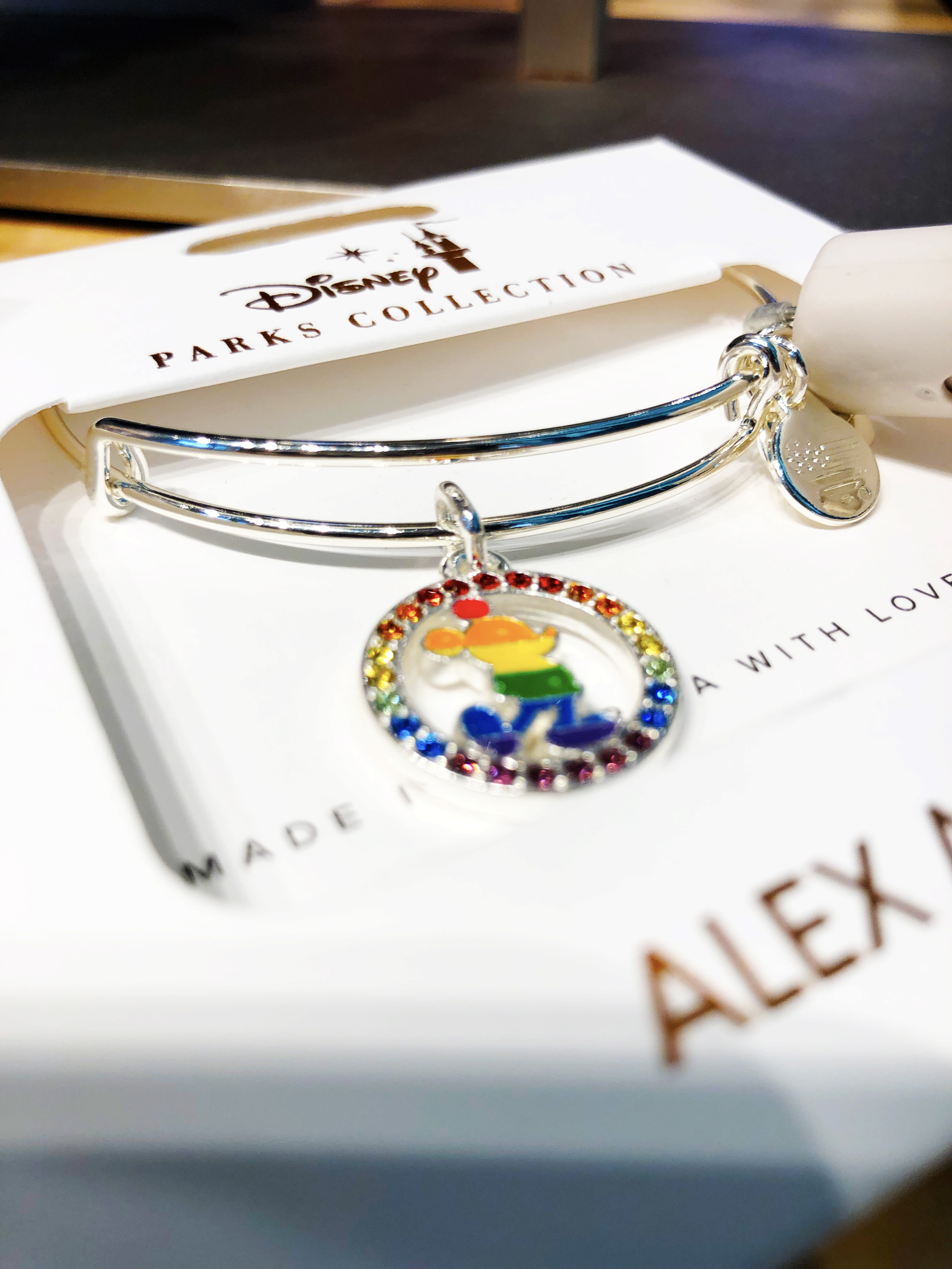 A left-angle shot of a packaged Alex and Ani bracelet. Bracelet is coated in silver with a circle charm featuring gems that circle into a rainbow with a full-body Mickey in a rainbow-striped pattern.
