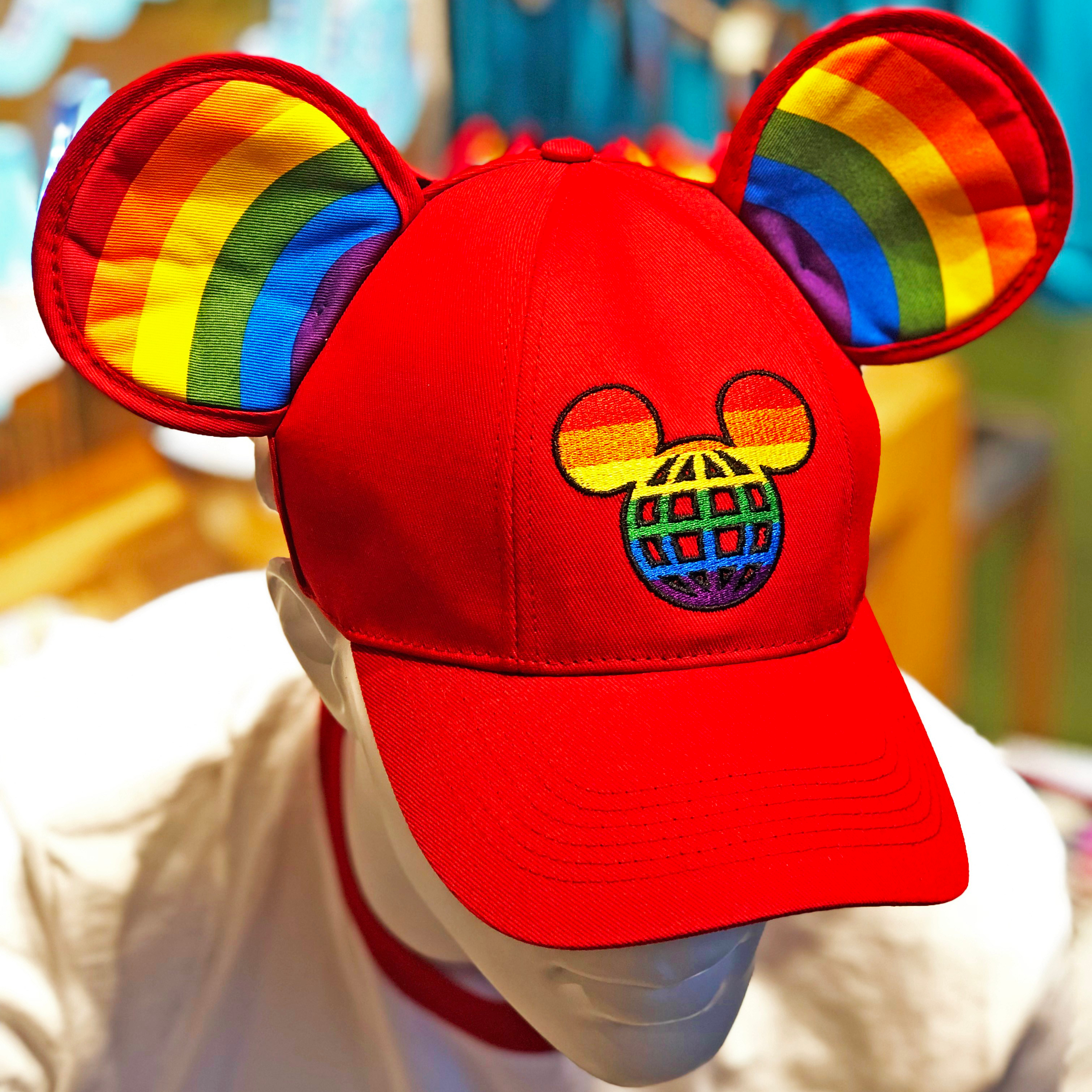 A zoomed-in photo of a baseball cap that has Mickey ears on it with rainbow-striping inside each year. Hat is red. In center of hat, it features a rainbow Mickey with a graph-like body. Hat is on mannequin.