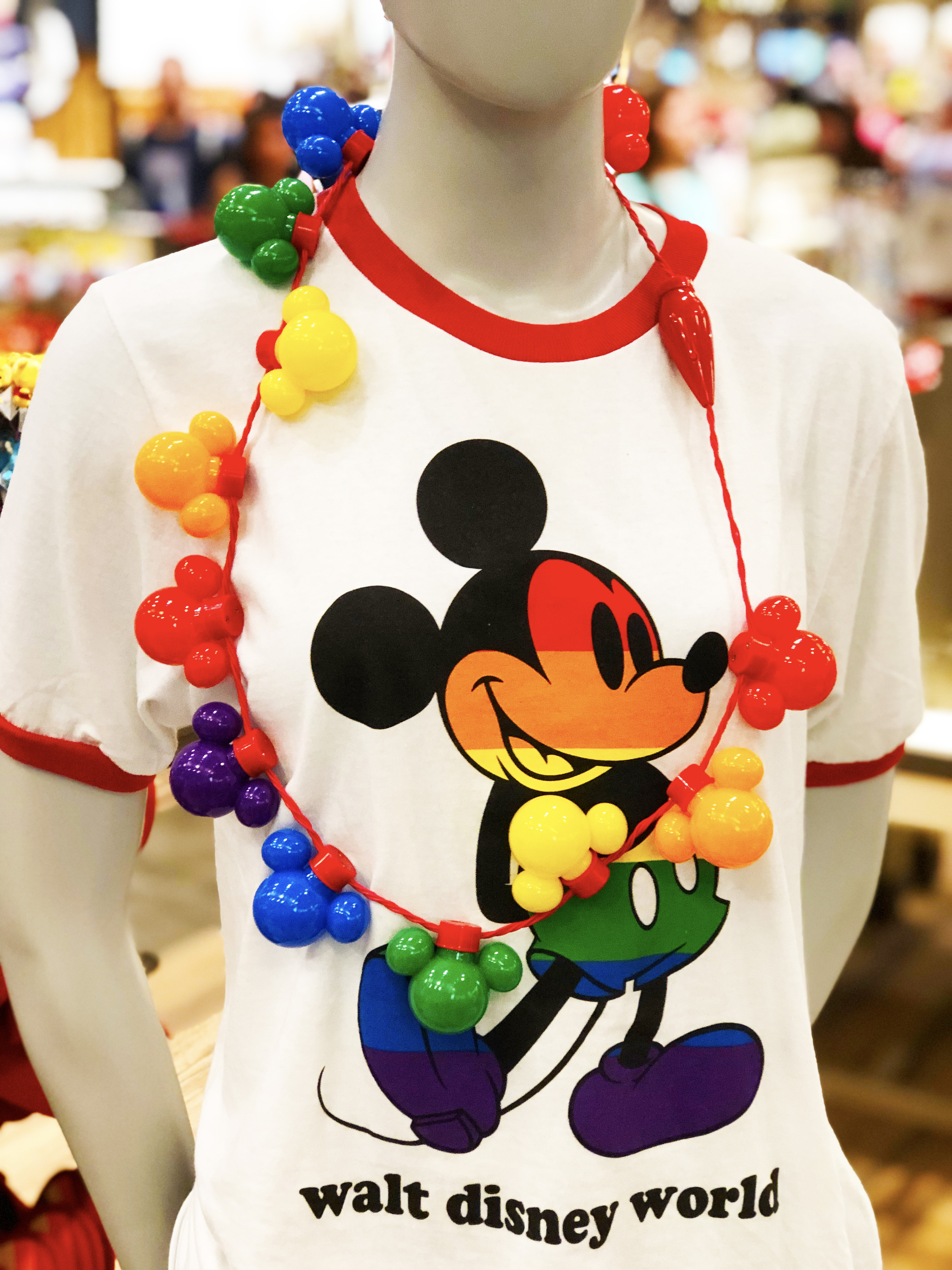 Mannequin decorated in light-up necklace with light-up Mickey heads that are each a different color to represent the rainbow on a red wire string. Mannequin is wearing the white tee with the classic Mickey sticking out his foot with his arms crossed behind his back (Mickey is rainbow-striped in the order from top to bottom of red, orange, yellow, green, blue, purple). Rounded collar and cut-off for the sleeves on the tee are red.