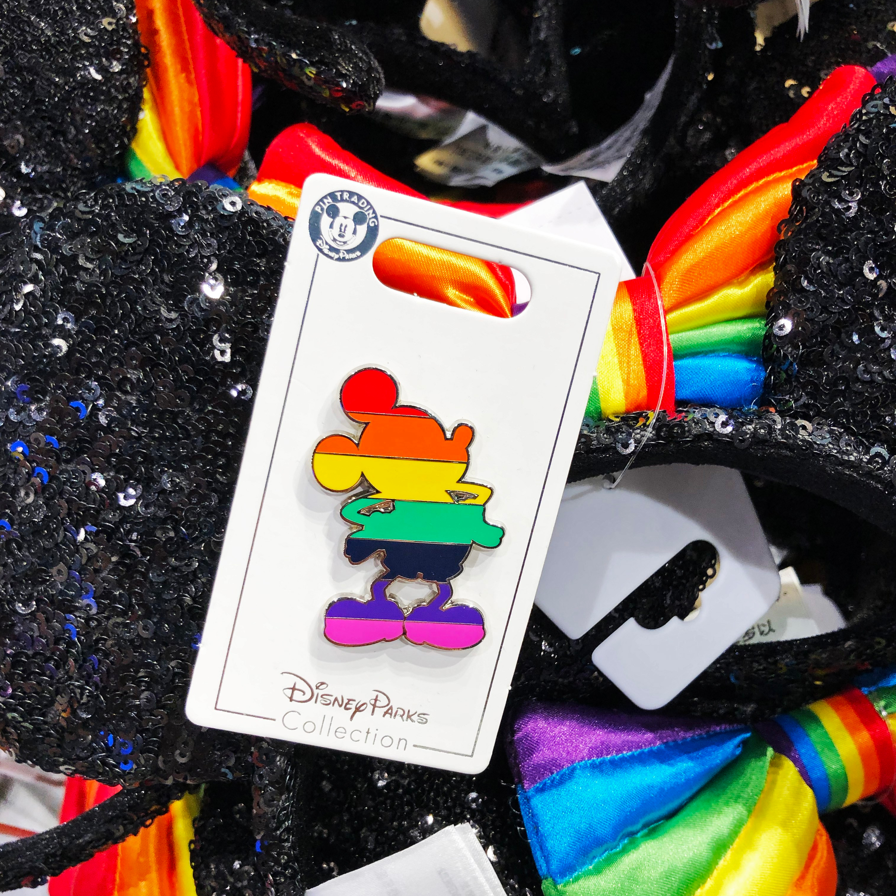 Rainbow-striped pin shaped of body of Mickey Mouse laying on top of black sequined Minnie ears with rainbow bow.
