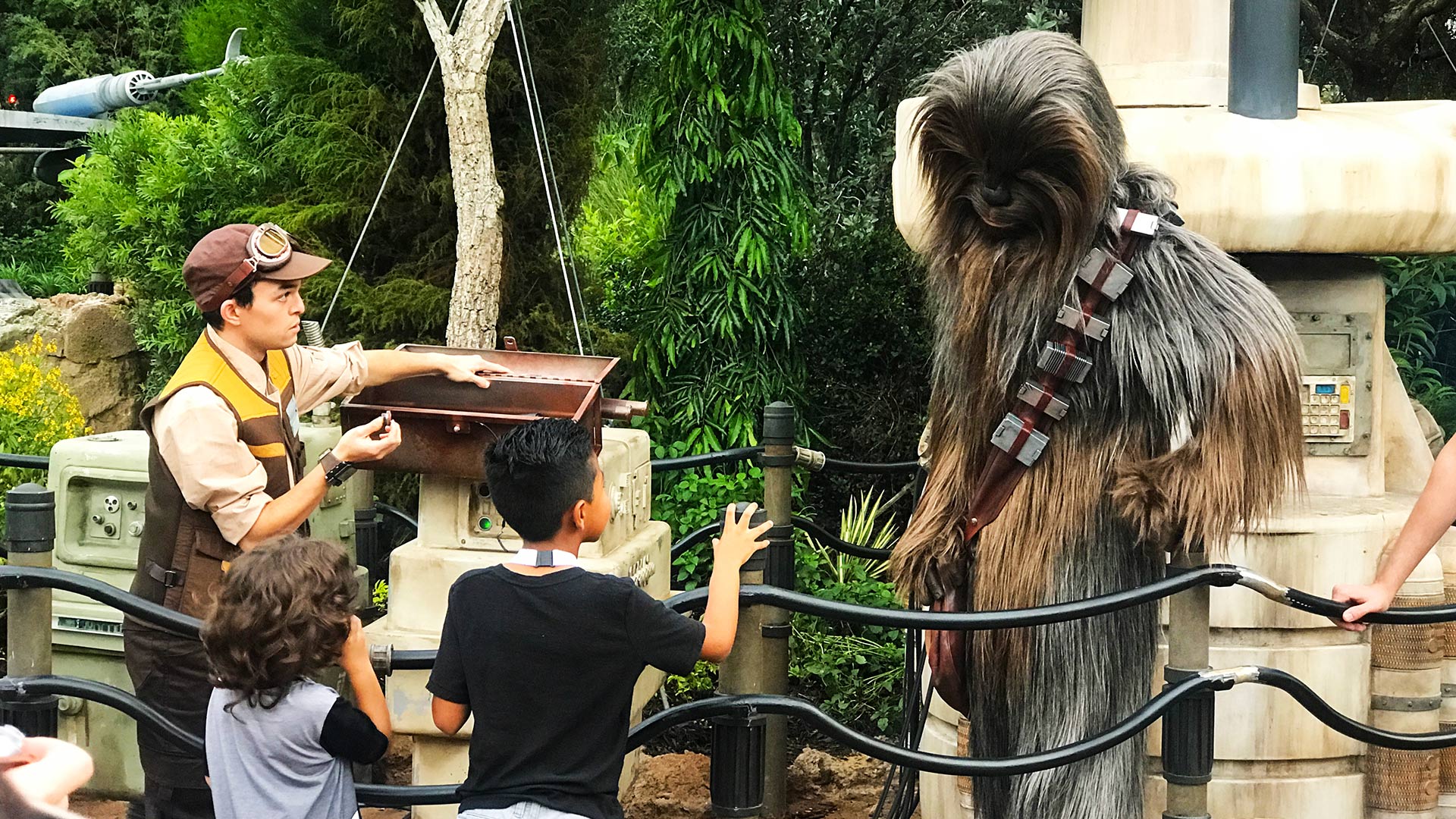Chewbacca (right) with hand on his hip as he looks down at two children (boys, bottom left of photo) speaking to him. Male cast member is looking at Chewbacca while next to the children (left) while holding something in his right hand. Daytime shot. Land of Batuu. Star Wars: Galaxy's Edge at Disney's Hollywood Studios®. Lake Buena Vista, Florida.