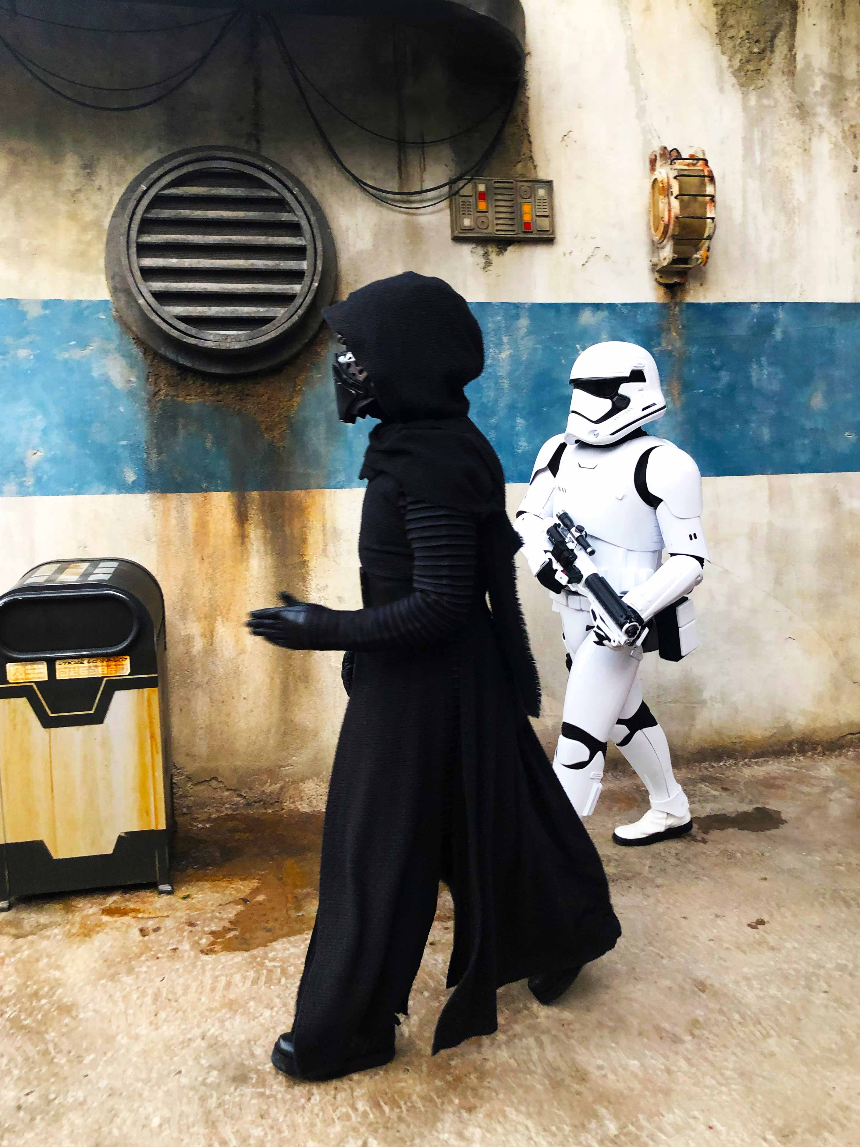 Daytime shot. Side view of Kylo Ren in full gear as he walks through Batuu with a Stormtrooper (to his right), armed with a trooper gun. Star Wars: Galaxy's Edge at Disney's Hollywood Studios. Lake Buena Vista, Florida.