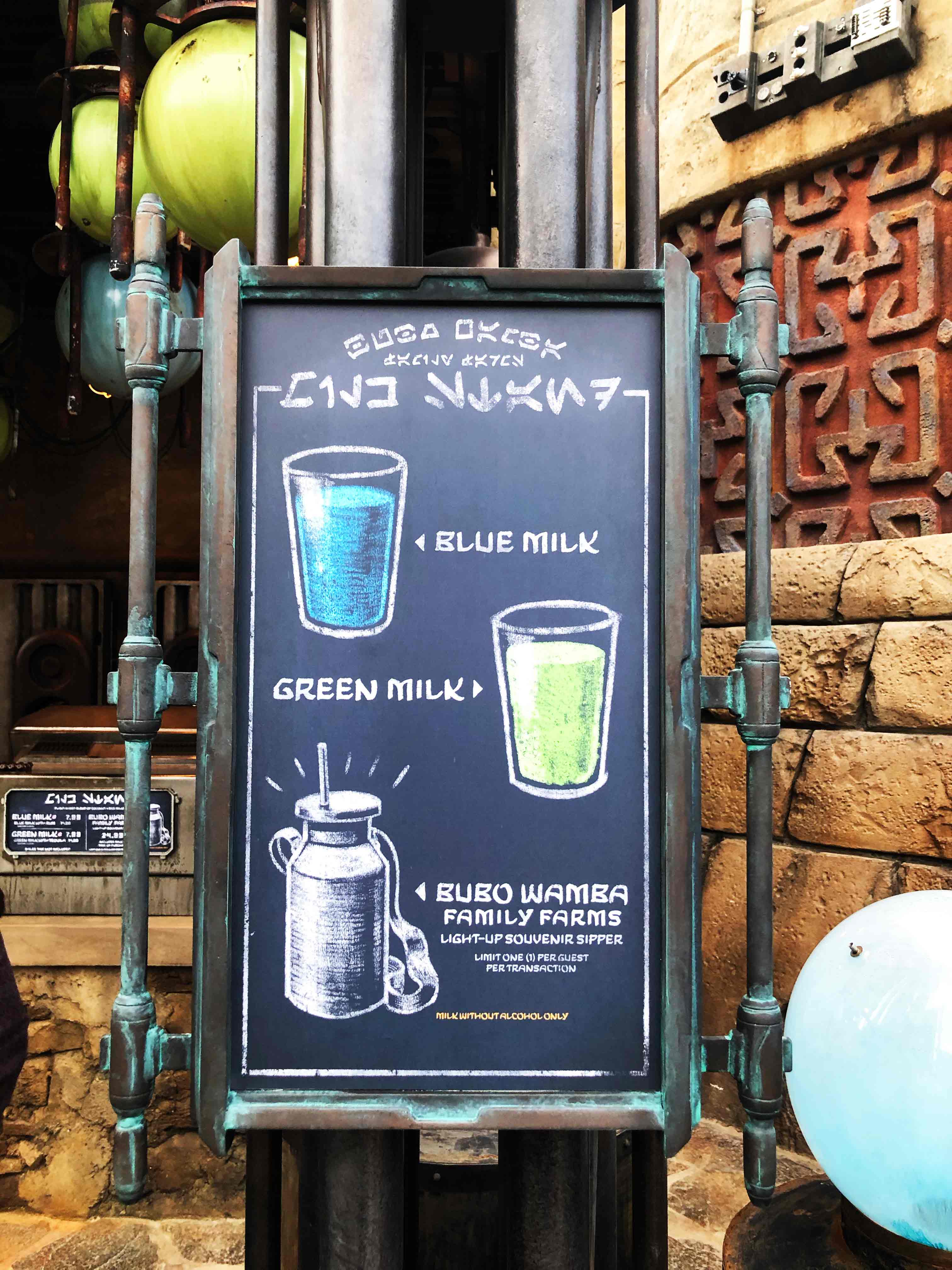 Chalkboard sign of milk options at The Milk Stand. To the right of the register counters. Daytime photo. In order from top to bottom, Blue Milk (text right, drawing of blue milk on left), Green Milk (text left, drawing of green milk on right), Bubo Wamba Family Farms Light-Up Souvenir Sipper (text right, drawing of sipper on left). Star Wars: Galaxy's Edge at Disney's Hollywood Studios®. Lake Buena Vista, Florida.