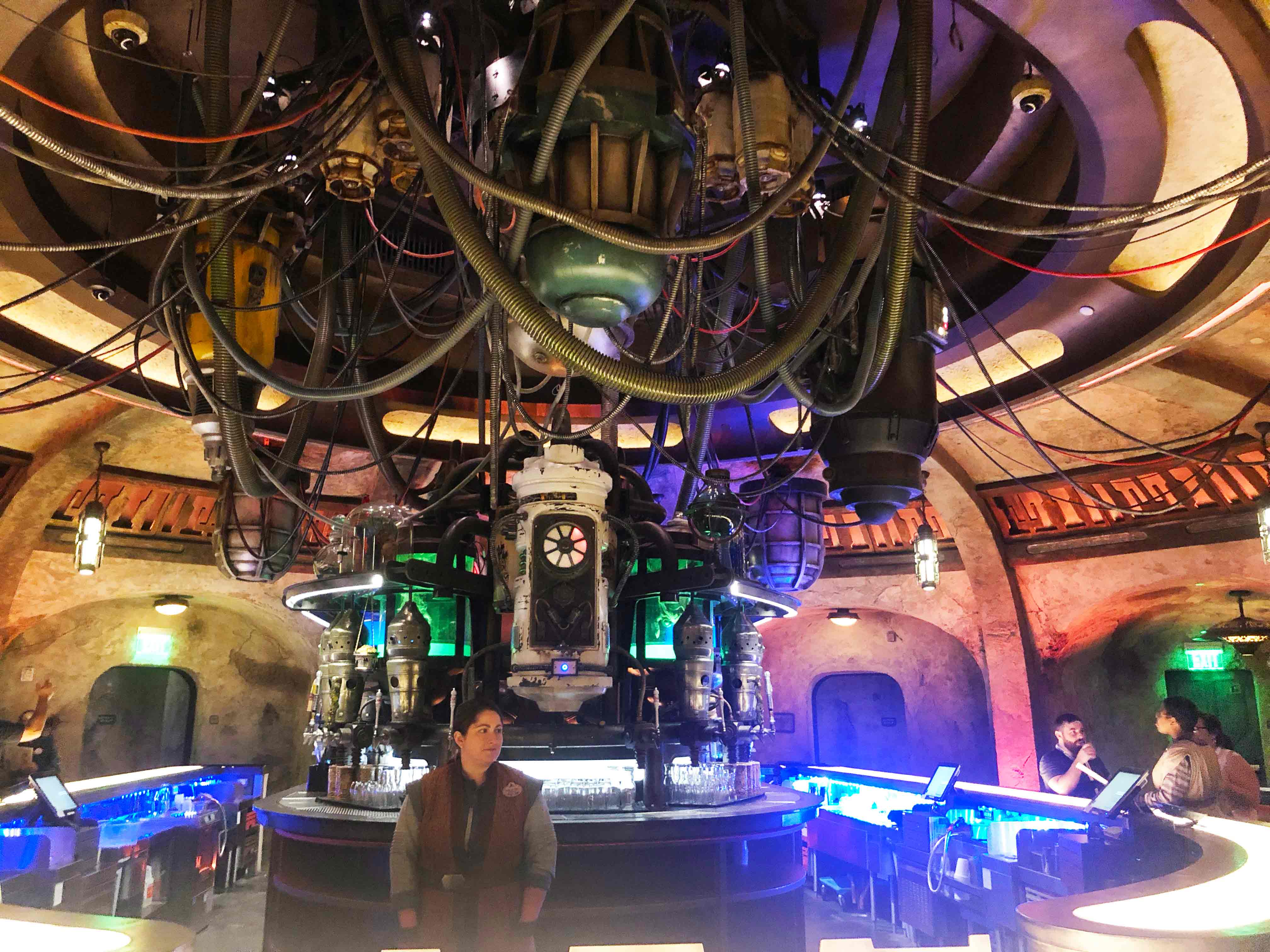 Photo of the bar inside of Oga's Cantina. A female cast member is standing behind the bar looking off to the right. Three guests sit at the right of the bar circle talking amongst each other. Star Wars: Galaxy's Edge at Disney's Hollywood Studios. Lake Buena Vista, Florida.
