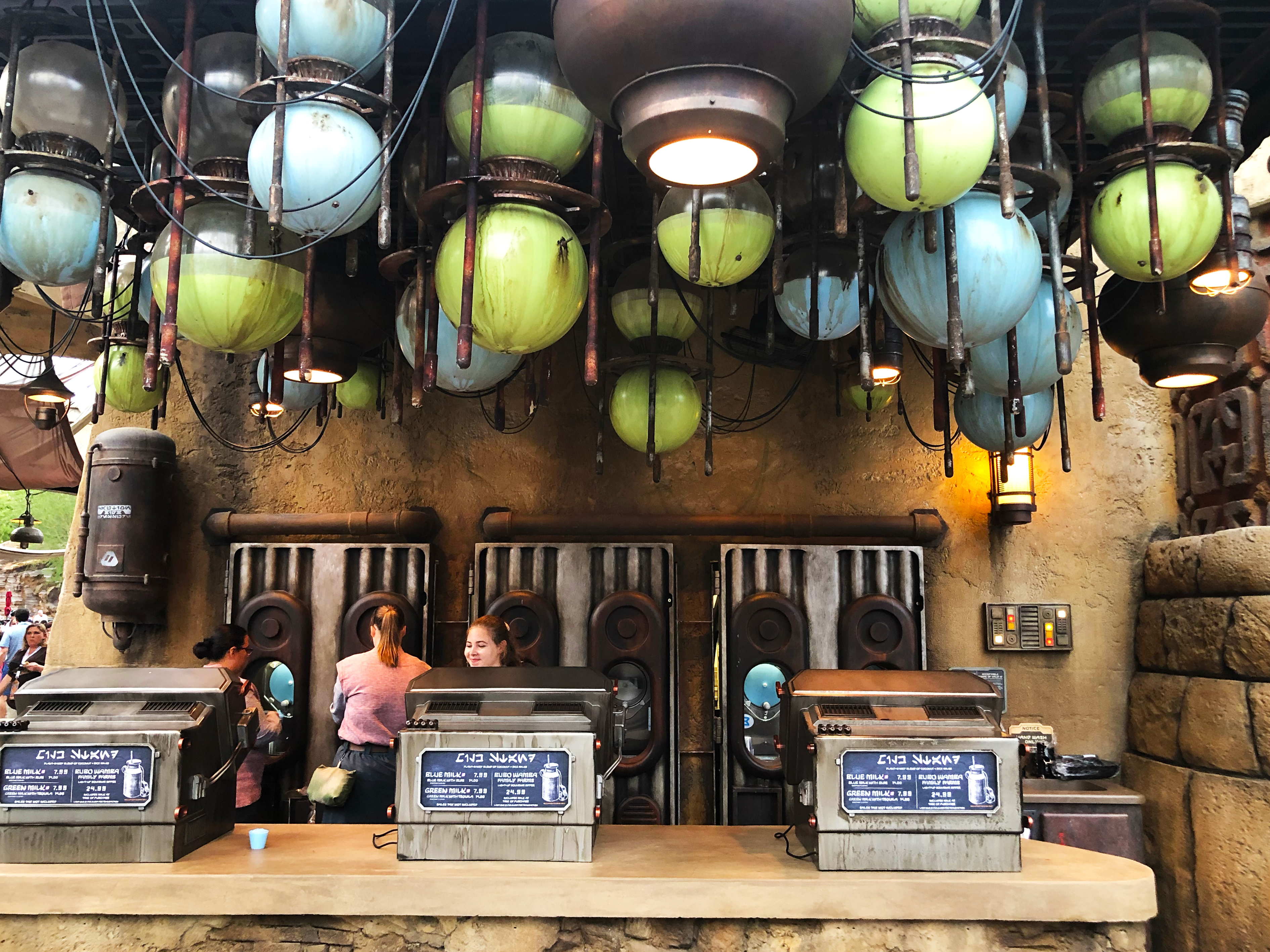 A daytime shot of The Milk Stand. Three cast members are seen behind the counter of cash registers, one (female, middle) is smiling while two (both females) have their backs turned as they dispense blue milk from the machines. Star Wars: Galaxy's Edge at Disney's Hollywood Studios®. Lake Buena Vista, Florida.