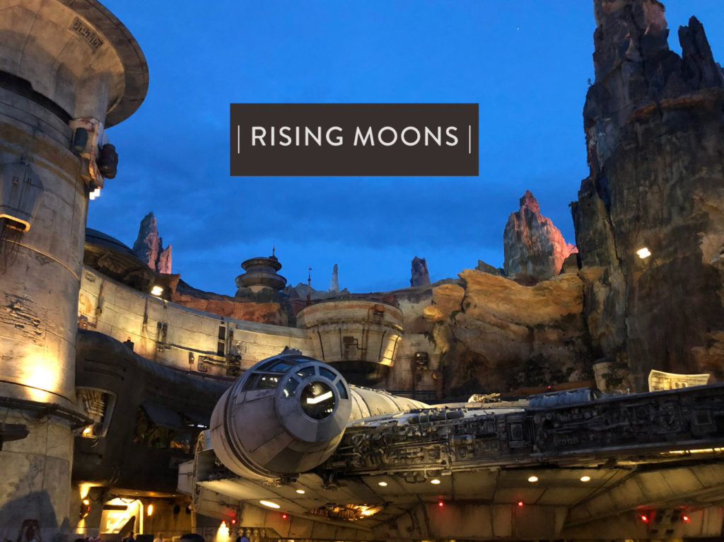 "Rising Moons" written in a boxed text over a photo of the Millennium Falcon during the evening. Outdoor shot. Star Wars: Galaxy's Edge at Disney's Hollywood Studios®, Lake Buena Vista, Florida.