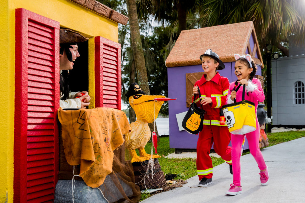 A boy (left) dressed as a fire fighter and a girl (right) dressed as a cat as they trick-or-treat to a man (far left) sticking his head out of a LEGO house dressed as a pirate during LEGOLAND Florida Resort's Brick or Treat.