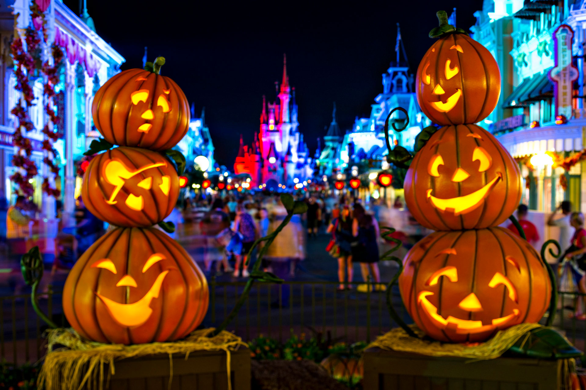 Two sets of 3-tiered glowing Halloween pumpkins with faces featuring the Cinderella Castle and Main Street U.S.A. in the distance (glowing in colors of purple, red, blue, and soft green) for Mickey's Not-So-Scary Halloween Party at the Magic Kingdom® Park. Nighttime shot. Lake Buena Vista, Florida.