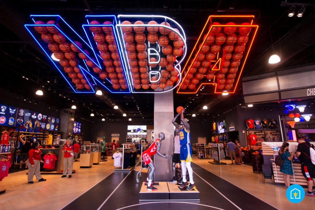 Inside of the entrance of NBA Experience at Disney Springs® (Lake Buena Vista, Florida). Giant "NBA" sign lit up in blue, white, and red with mannequins underneath holding basketballs.