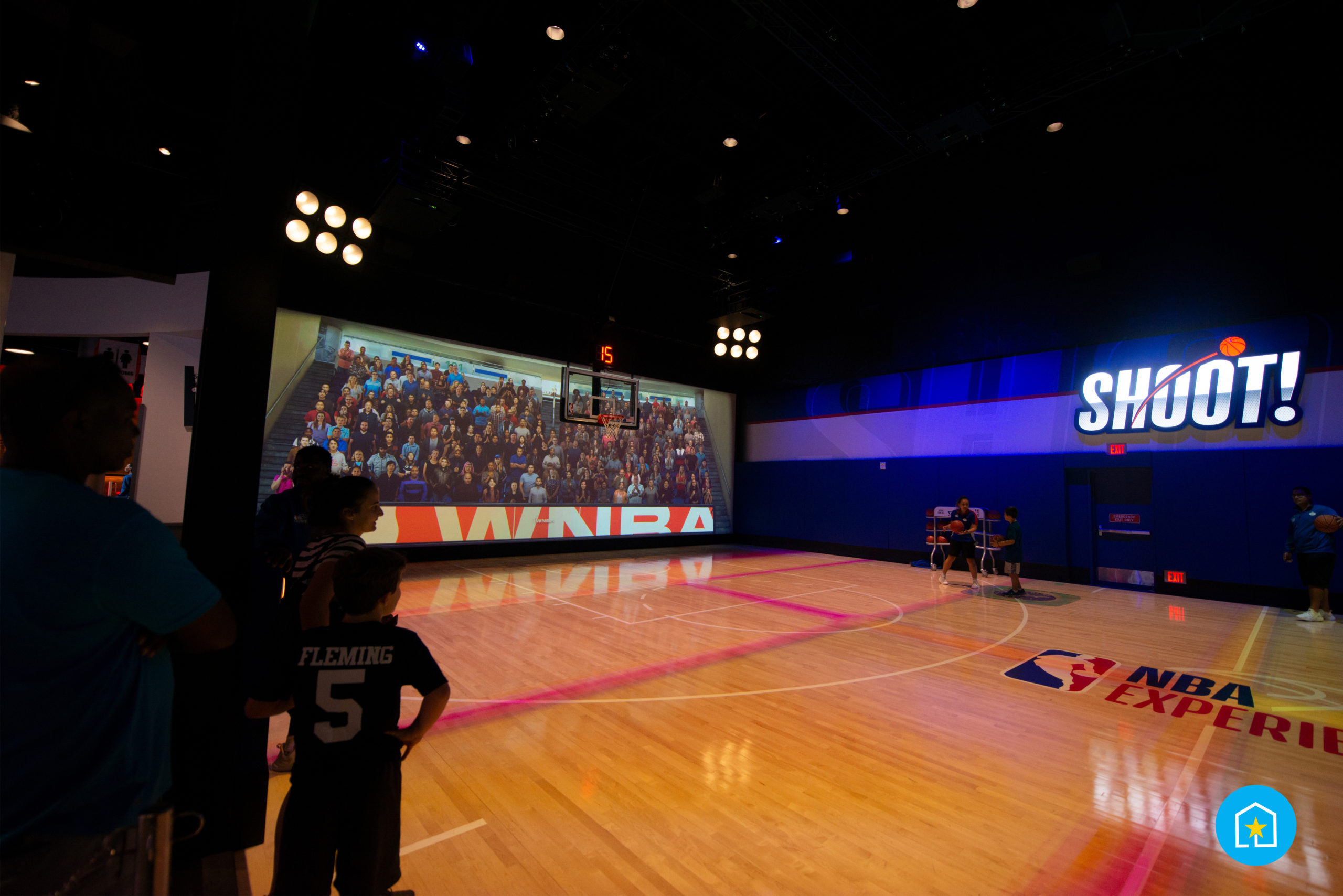 Basketball court inside of the NBA Experience in Disney Springs® (Lake Buena Vista, Florida). Wall on left features giant projection of crowd on bleachers cheering, wall to the right says "SHOOT" to signify the particular room. Left of photo shows backs of guests waiting to enter the court as they watch two individuals on the court, one, man (left), holding a basketball and explaining how to play to boy (right).