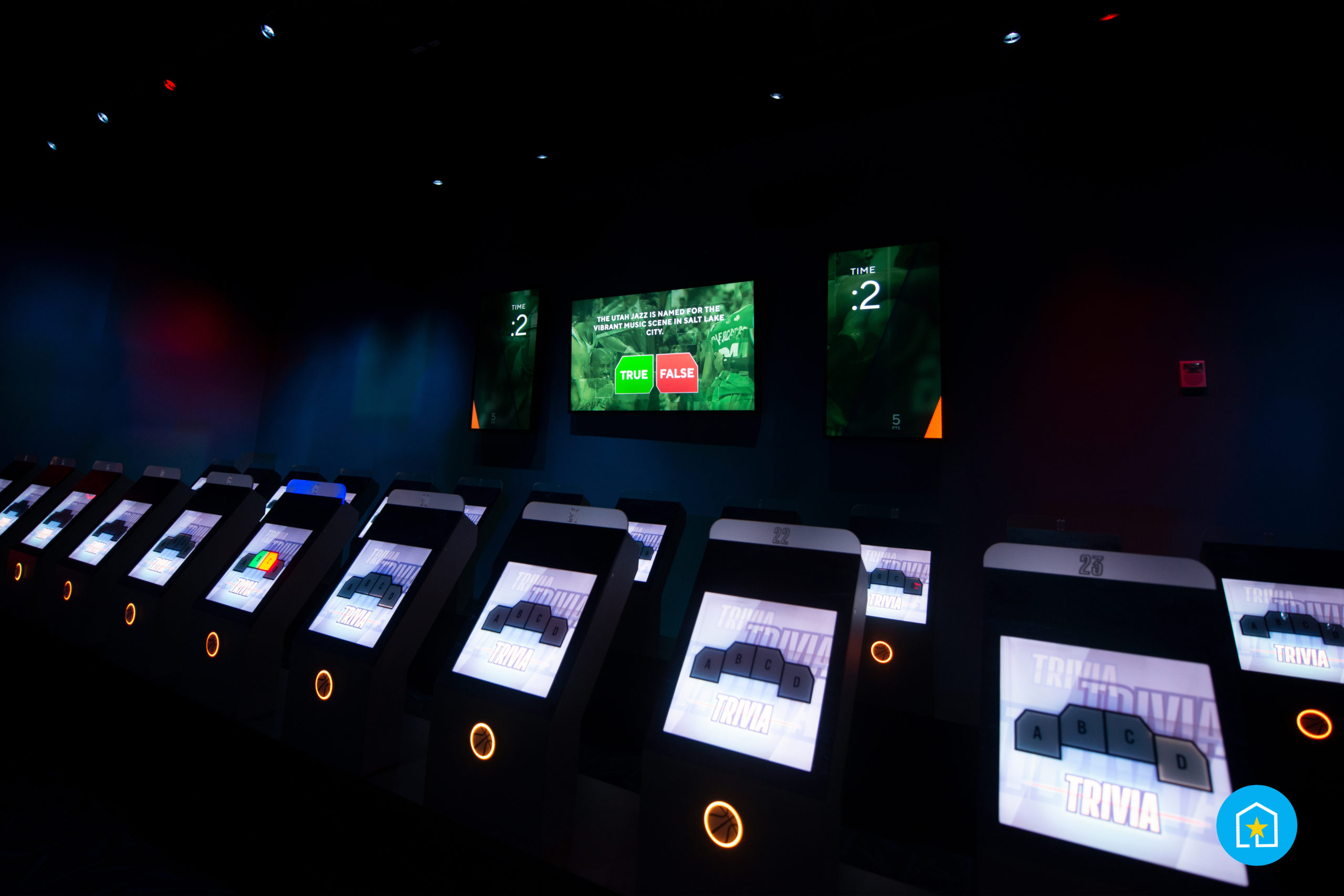 Low-lit room for NBA Trivia. Seen in photo, giant screen showcasing 2 seconds left on the clock to answer a trivia question. Smaller screens with Magic Band tap circles lined up across the room in two separate rows.