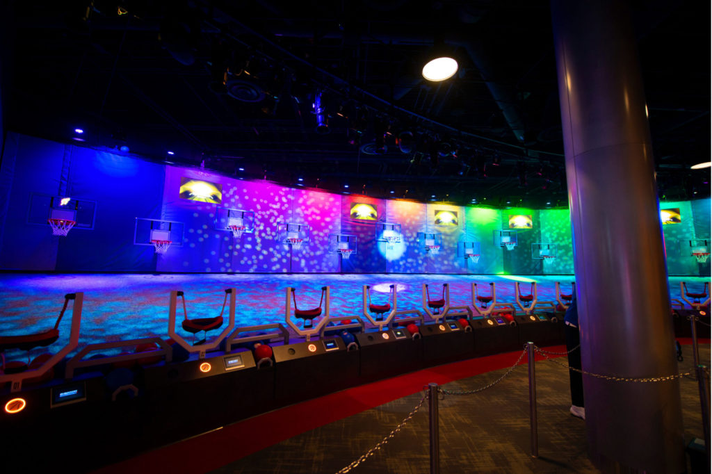 View from queue for Slingshot inside of the NBA Experience at Disney Springs® (Lake Buena Vista, Florida). View shows multiple basketball slingshots and wall behind them is decorated in hues of multicolored lights.