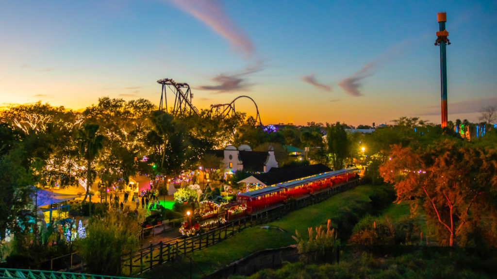 A distant shot of a sunset across Busch Gardens Tampa Bay, featuring some rollercoasters and a holiday train on the outskirt of the park. For Busch Gardens Christmas Town.