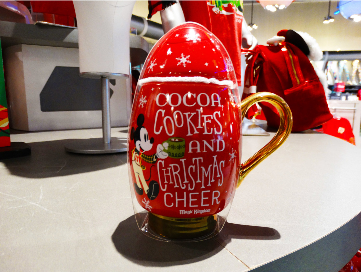 Front of Mickey's Very Merry Christmas Party Mug on shelf, reading "Cocoa, Cookies, And Christmas Cheer, Magic Kingdom®" with Mickey Mouse holding a cup of cocoa on the left of the text, snowflakes all over the mug. Lake Buena Vista, Florida.
