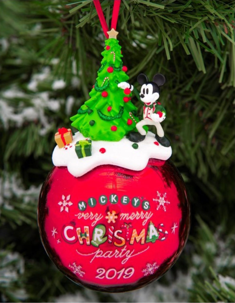 Mickey's Very Merry Christmas Party 2019 limited release red Christmas ornament hanging on tree. Close-up shot. Ornament features Christmas tree on top surrounded by presents and snow with classic Mickey mouse on the right side decorating the tree. The main ornament is round and features snowflakes and the name of the event printed on the front. The Magic Kingdom® Park. Lake Buena Vista, Florida.