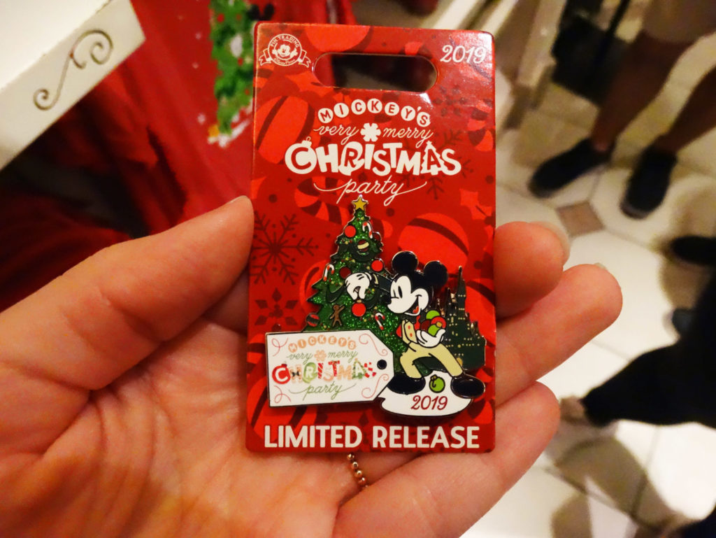 Hand holding limited release pin from Mickey's Very Merry Christmas Party 2019 at the Magic Kingdom® Park. Pin design features classic Mickey Mouse decorating a Christmas tree next to a large gift tag that reads the name of the event. 2019 printed under Mickey's feet. Cinderella castle behind Mickey Mouse traced in gold and colored in dark, almost black-like color. Lake Buena Vista, Florida.
