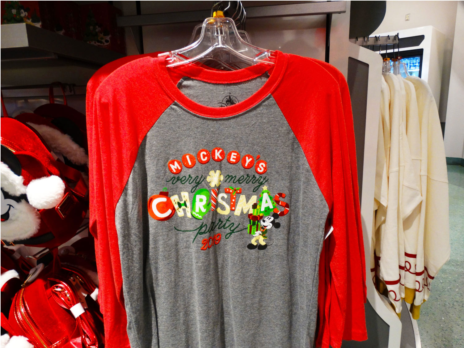 Photo of long sleeve tee hanging inside of a store at Mickey's Very Merry Christmas Party 2019 at the Magic Kingdom® Park, grey with red sleeves featuring the name of the party in Christmas Ornament, Light, and Classic Mickey graphic on center. Lake Buena Vista, Florida.