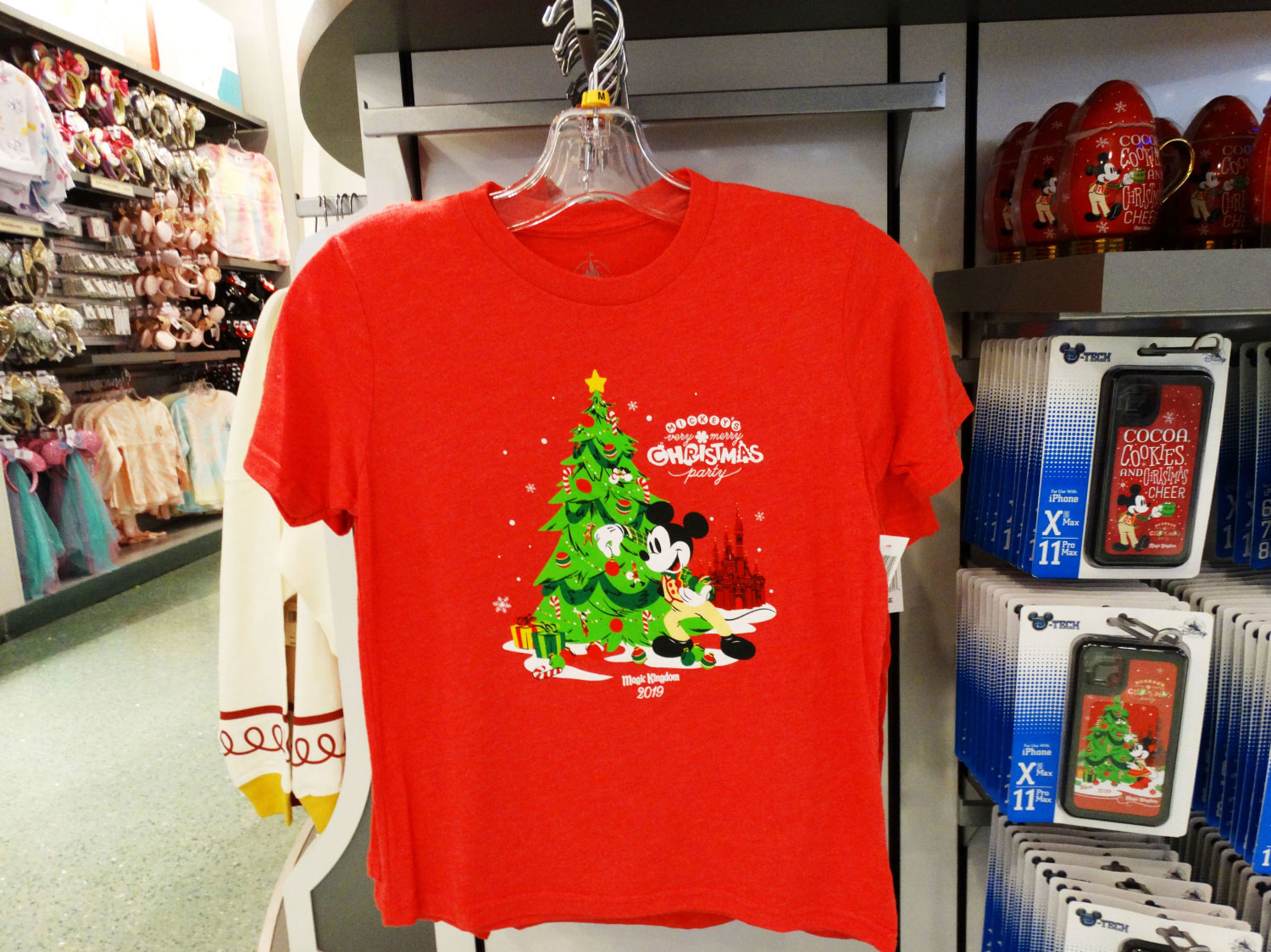 Red short sleeve tee on a hanger inside of a store during Mickey's Very Merry Christmas Party 2019 at the Magic Kingdom® Park. Shirt sports Christmas Tree graphic with Mickey Mouse decorating the tree and the name of the event on the top right of the center graphic. Lake Buena Vista, Florida.