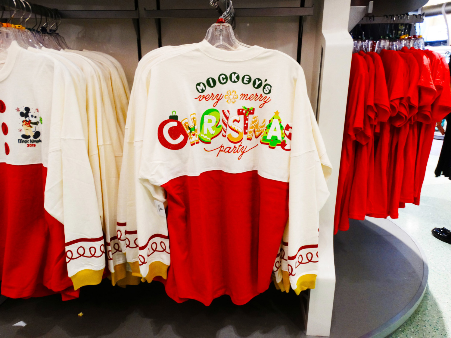 Limited edition Mickey's Very Merry Christmas Party Spirit Jersey hanging on rack in-store at the Magic Kingdom® Park. Name of the event printed on the top half of Jersey with cream white background and Christmas graphics, the bottom half of jersey is red. Lake Buena Vista, Florida.