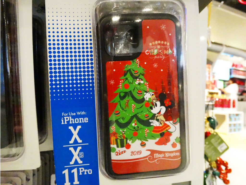 Limited Release iPhone case for Mickey's Very Merry Christmas Party 2019 at the Magic Kingdom® Park in Lake Buena Vista, Florida. Case is hanging on rack and features a red background decorated in snowflakes and the name of the event on the top right. Main graphic features classic Minnie Mouse (right) decorating a Christmas tree (left) with ornaments as they are both surrounded with snow, presents, ornaments, and a candy cane. Underneath main graphic are is "2019" and "Magic Kingdom".