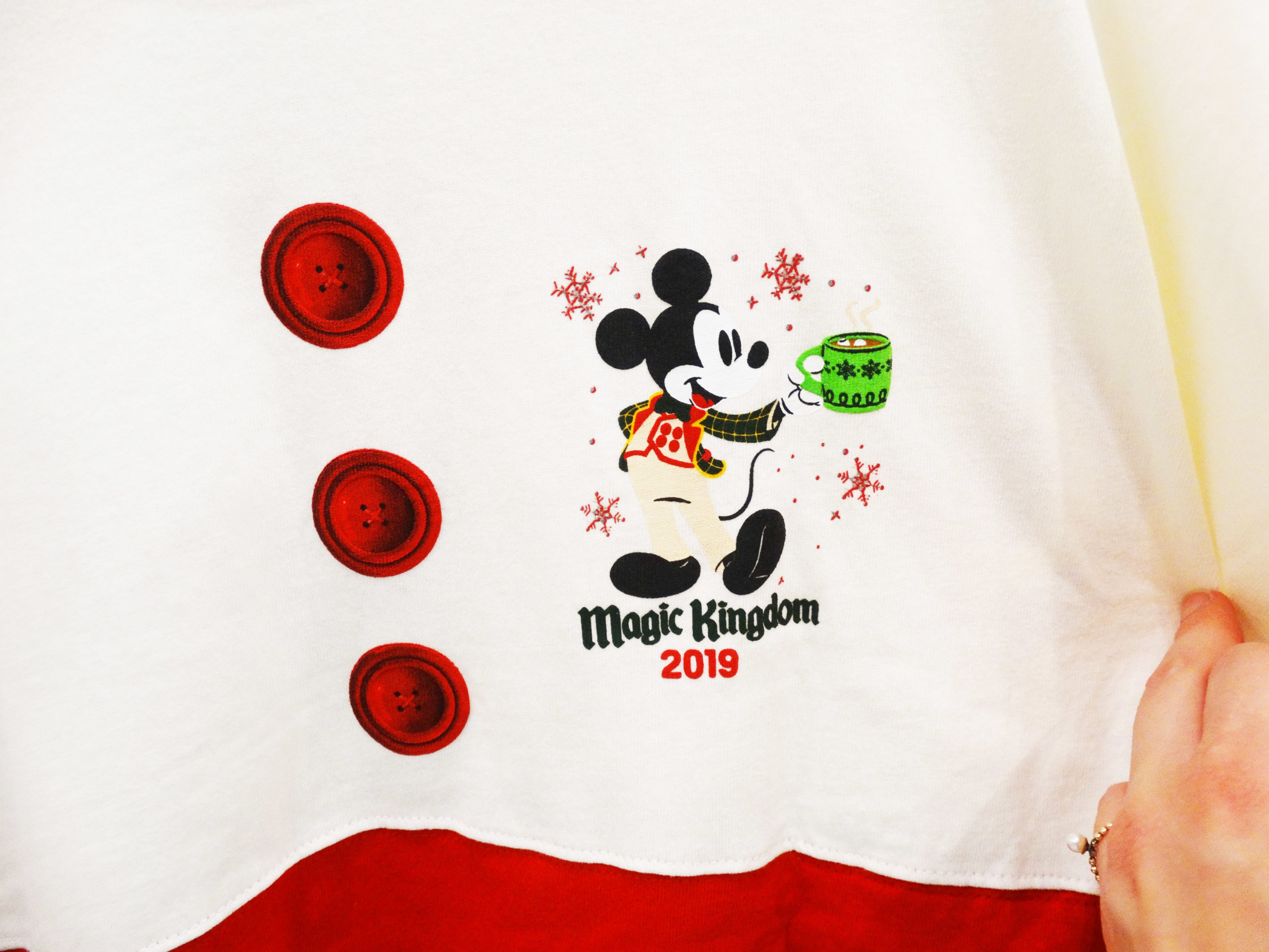 Close up of the front of the Mickey's Very Merry Christmas Party 2019 Spirit Jersey. 3 red button graphic on cream-colored fabric with Mickey Mouse holding a cup of cocoa over the words "Magic Kingdom 2019". The Magic Kingdom® Park, Lake Buena Vista, Florida.