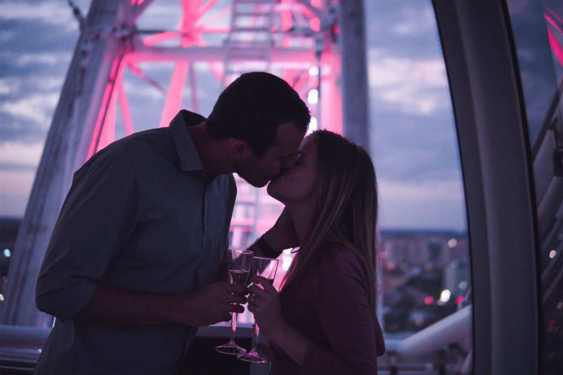 Dim-lit sunset photo of couple mid-kiss while holding two champagne glasses at the top of The Wheel at ICON Park, International Drive, Orlando, Florida.