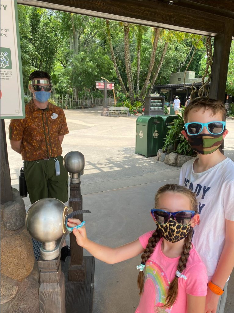 Cast member (left corner) watching two kids (little girl with her Magic Band on the Mickey scanner and little boy behind her) as they all look at the camera before entering Disney's Animal Kingdom® Theme Park. All people are wearing face masks and sunglasses. Cast member is also wearing face shield. Daytime shot. Lake Buena Vista, Florida.