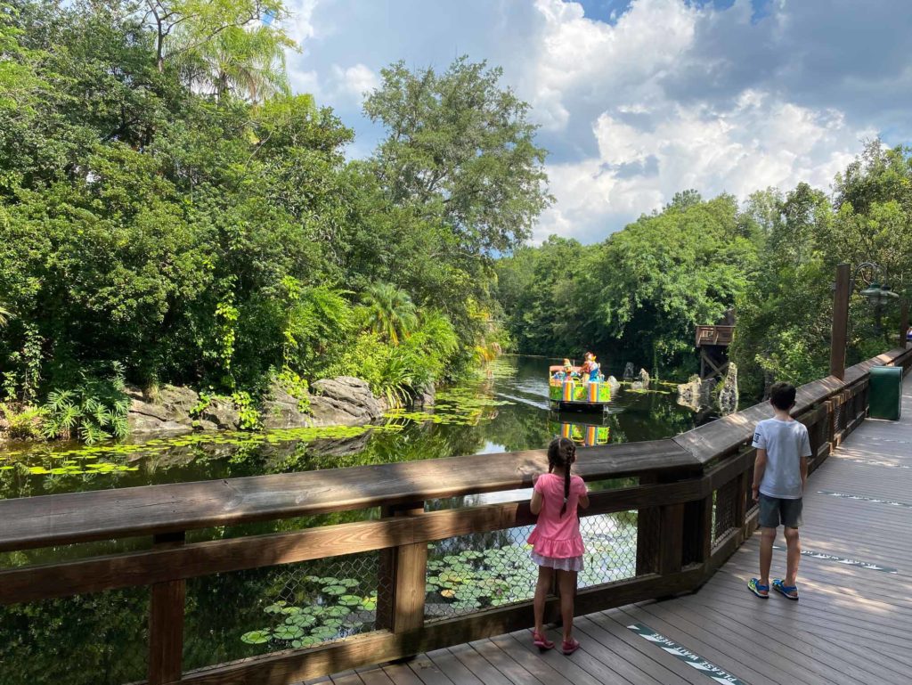 Girl (left) and boy (right) look over a wooden railing out to the lake where a water float boat cruises through with Daisy and Donald Duck at Disney's Animal Kingdom® Theme Park. Daytime shot.