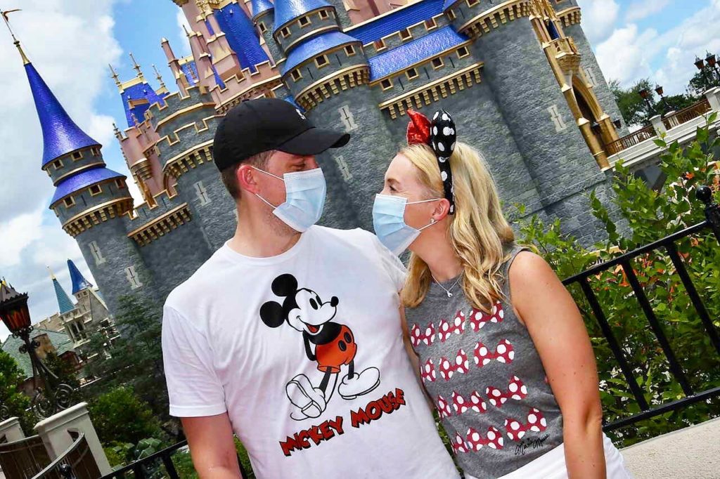 Daytime photo of man (left) and woman (right) as they look at each other (both wearing face masks) posed in front of Cinderella Castle at the Magic Kingdom® Park. 2020. Photographed at an angle. Lake Buena Vista, Florida.