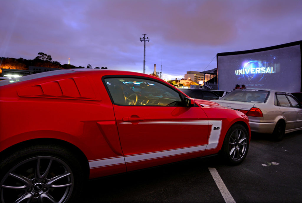 A sunset photo of a red Mustang at a parking lot for the Halloween Drive-In at Old Town Kissimmee. Right side of car. Facing direction of projector screen that features a projection of the "Universal" logo as a movie begins. 2020.
