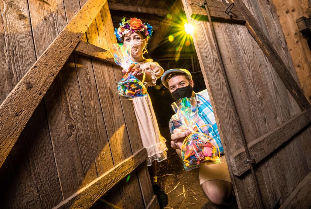 Female (left) and male (right) dressed in costume in a barn holding goodie bags of Halloween candy for The Haunted Road's daytime family-friendly drive-in event. 2020. Orlando, Florida.