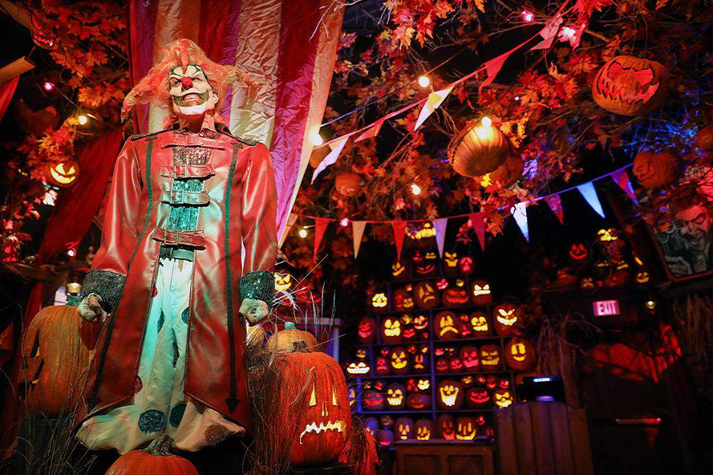 Photo of inside the Halloween Horror Nights Tribute Store at Universal Studios Florida, 2020. A lifelike clown figure stands smiling as he is surrounded by pumpkins, an artificial tree, and red and orange lighting. Orlando, Florida.