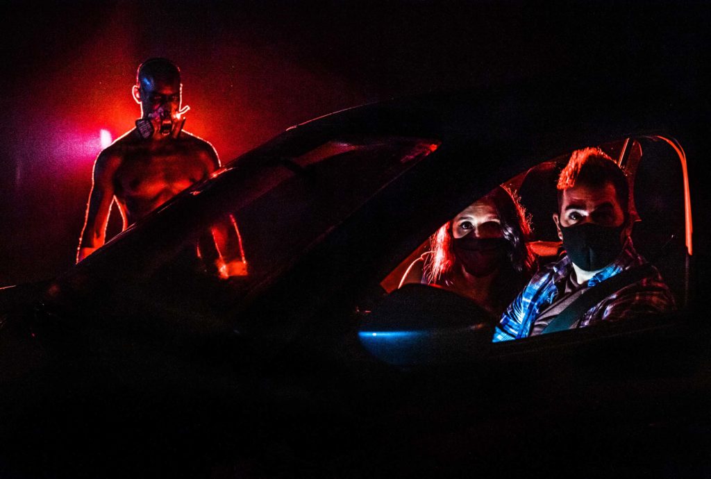 Nighttime photo of female (passenger seat) and male (driver seat) wearing face masks with fearful eyes as a scareactor approaches the passenger side of the vehicle with a red glow behind him. The Haunted Road. Orlando, Florida, 2020.